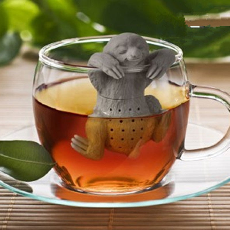 Gray sloth-shaped tea infuser hanging over the rim of a clear glass mug filled with amber tea